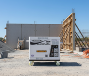 A Paddock Diesel Inverter 5kW Generator stands on a gravel construction site before a partially built concrete wall with exposed rebar and wooden frames, all under a clear blue sky.