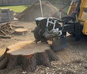 A Paddock Swinging Stump Grinder Attachment - Suits STRKL1200 is grinding a large stump in a backyard, scattering wood chips and debris, with piles of soil, cut branches, and pavers visible in the background.