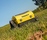 A Paddock Remote Controlled Mower 7.5HP - 530mm, in yellow, drives over green grass beneath a blue sky with trees in the background.