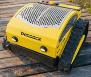 A yellow Paddock Remote Controlled Mower 7.5HP - 530mm with tracks sits outdoors on wooden planks, featuring a perforated cover, top control buttons, and a powerful petrol engine.