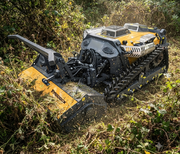 A Paddock™ Remote Controlled Mower/Slasher/Mulcher 35HP - 1150mm, featuring a durable yellow and black body, trims thick vegetation in a dense, overgrown area. This Paddock slope mower is equipped with a robust track drive system, enabling it to navigate challenging terrain with ease.