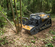 The Paddock™ Remote Controlled Mower/Slasher/Mulcher 35HP - 1150mm by Paddock mulches small trees and brush on a forest floor, surrounded by greenery and scattered branches.