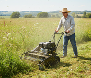 A person in a green shirt and gloves operates the Paddock Walk Behind Flail Mower by Paddock to cut tall grass for vegetation control in a sunny field bordered by trees and a wooden fence.