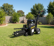 A Paddock Walk Behind Trencher with Guide Wheel by Paddock sits on a neatly mowed lawn in a fenced backyard, surrounded by trees and shrubs on a sunny day.