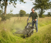 A man wearing ear protection uses a Paddock Walk Behind Tractor Flail Mower Attachment by Paddock to cut tall grass in a rural field surrounded by trees under a partly cloudy sky.