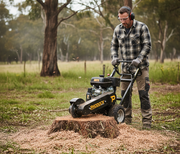 Wearing safety gear, a man operates a Paddock Commercial Stump Grinder by Paddock to remove an outdoor tree stump, surrounded by grass, trees, and scattered wood chips.