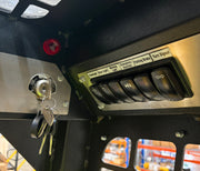 Close-up of the Paddock Tracked Cabin Loader Pro control panel, showing keys in the ignition and labeled switches for headlights, hazard lights, parking brake, and turn signal. Metal grate and warehouse shelving are visible in the background.