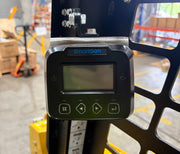 A close-up of a Paddock SmartGen digital control panel with screen and four buttons, mounted on a Paddock Tracked Cabin Loader Pro featuring a 40hp Perkins turbo diesel engine, in an industrial warehouse with shelves and boxes.