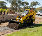 A Paddock Tracked Mini Loader Pro (40hp Perkins Turbo Diesel) transports soil in its bucket at a construction site, with stone landscaping, green grass, and trees in the background.