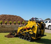The Paddock Tracked Mini Loader Pro with a 40hp Perkins Turbo Diesel engine moves soil on a grassy lawn, with mulch, shrubs, and a white pickup in the background—ideal for landscaping under a clear blue sky.