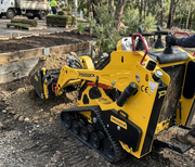 A Paddock Tracked Mini Loader Pro with a 40hp Perkins Turbo Diesel Engine moves soil at an outdoor landscaping site, demonstrating strong construction capability as workers and a truck can be seen in the background among trees and garden beds.
