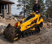 A person operates a yellow Paddock™ Tracked Mini Loader Elite, powered by a 58hp Yanmar 2.1L 4-cylinder CRDi Turbo Diesel Engine, moving dirt at a construction site with rocks, soil, and trees in the background.