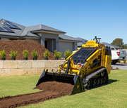 The Paddock Tracked Mini Loader (35hp Kubota Diesel) with Free 4in1 Bucket spreads soil on a lawn in front of a modern home featuring solar panels, stone retaining wall, potted plants, and a mulch pile beneath a clear blue sky.