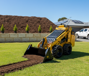 A Paddock Wheeled Mini Loader (35hp Kubota diesel, free 4in1 bucket) spreads soil on a mowed lawn in a landscaped yard with mulch pile, young trees, and a solar-paneled house in the background.