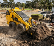 A Paddock Wheeled Mini Loader (35hp Kubota Diesel Engine) with Free 4in1 Bucket Bundle moves soil on a construction site, with dirt, gravel, stacked supplies, and trees in the background.